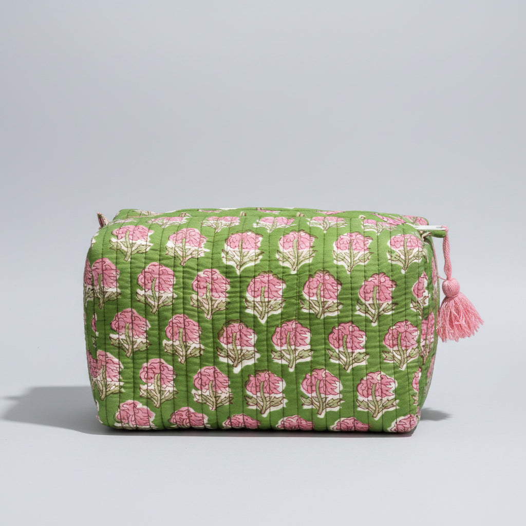 A green quilted cosmetic bag with a pink and white floral block print pattern, resting on a dark wooden surface against a dark gray textured background.