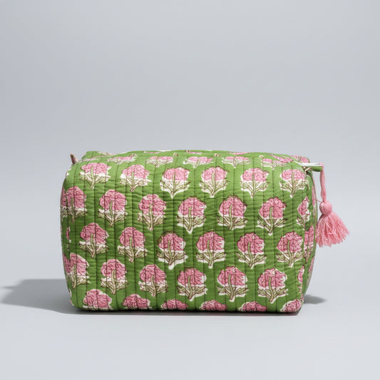 A green quilted cosmetic bag with a pink and white floral block print pattern, resting on a dark wooden surface against a dark gray textured background.