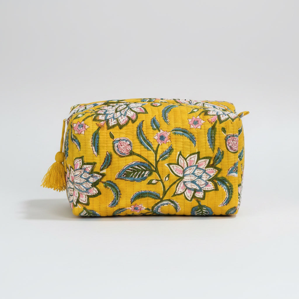 A rectangular yellow fabric pouch with a floral pattern in pink, green, and white, featuring a yellow tassel zipper pull, resting on a dark wooden surface against a dark gray background.
