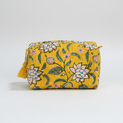 A rectangular yellow fabric pouch with a floral pattern in pink, green, and white, featuring a yellow tassel zipper pull, resting on a dark wooden surface against a dark gray background.