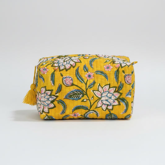 A rectangular yellow fabric pouch with a floral pattern in pink, green, and white, featuring a yellow tassel zipper pull, resting on a dark wooden surface against a dark gray background.