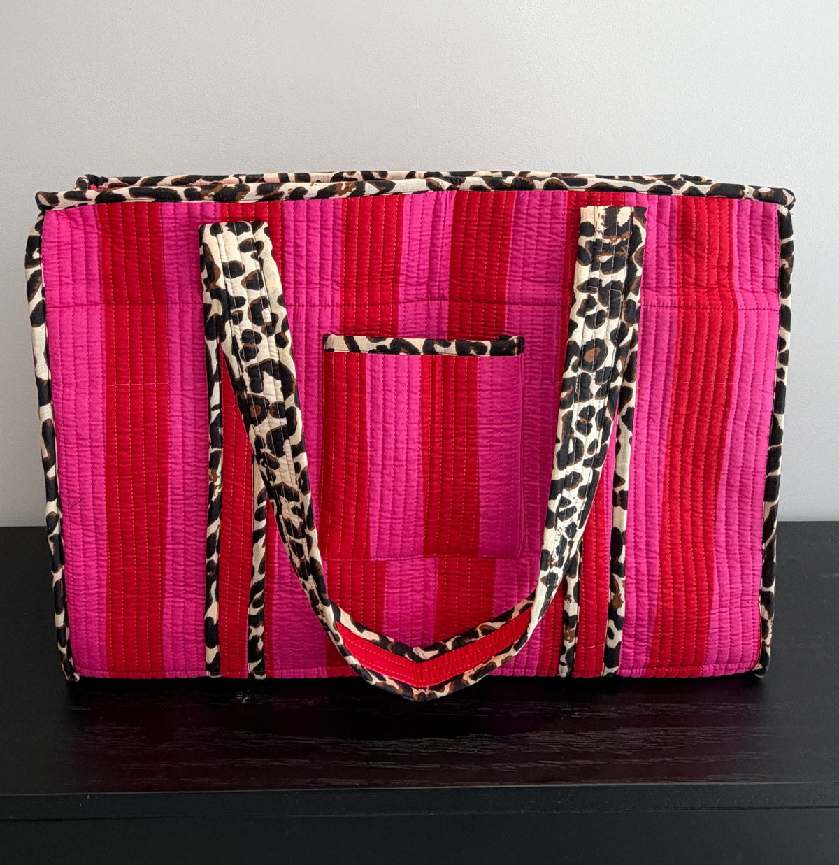 A quilted tote bag featuring vibrant pink and magenta striped panels, leopard print trim, and matching shoulder straps, displayed on a dark wooden surface against a plain light gray wall.