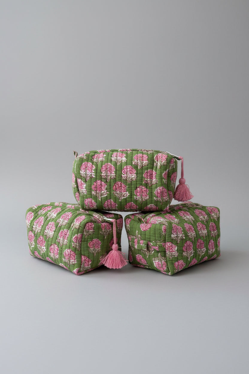 Three quilted cosmetic pouches stacked on a white marble surface, featuring a green and pink floral block print with pink tassels on the zipper pulls.