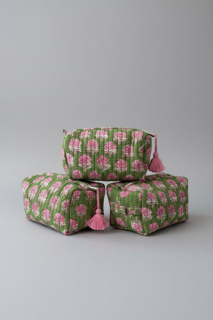 Three quilted cosmetic pouches stacked on a white marble surface, featuring a green and pink floral block print with pink tassels on the zipper pulls.