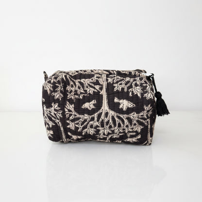 A black and beige quilted cosmetic pouch with a tree of life and bird pattern, featuring a black tassel zipper pull, set on a white surface with a blurred living room background.