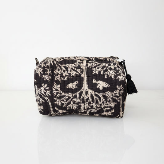 A black and beige quilted cosmetic pouch with a tree of life and bird pattern, featuring a black tassel zipper pull, set on a white surface with a blurred living room background.