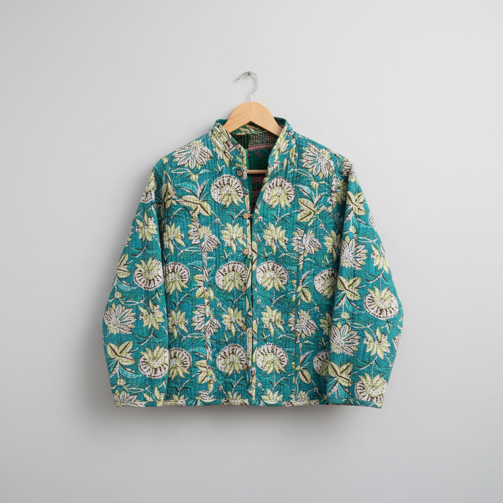 A teal quilted jacket with a cream and olive floral pattern, hanging on a wooden hanger against a dark wooden fence.
